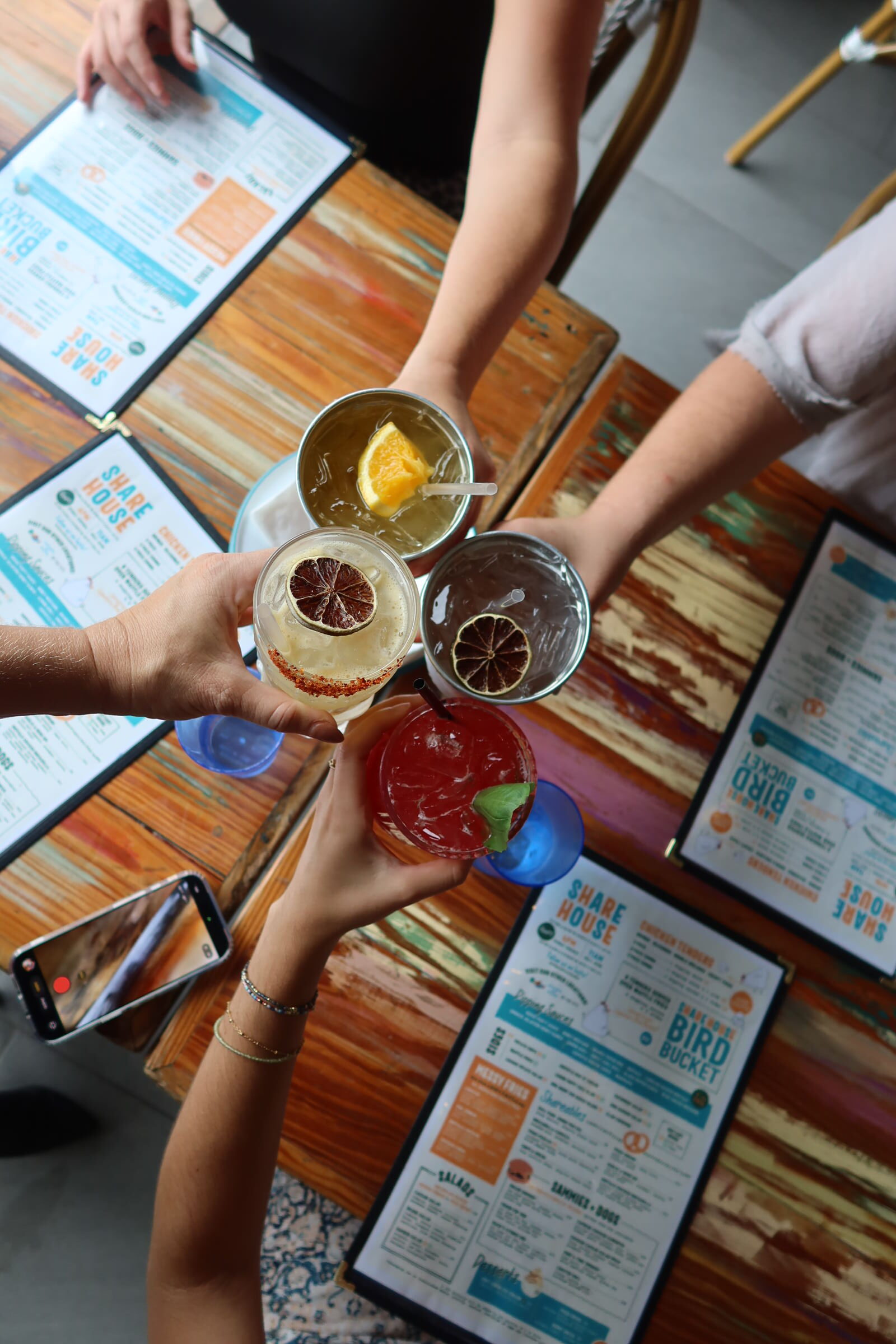 Overhead view of four women toasting colorful cocktails at Share House in Charleston, SC during a bachelorette party, with Share House menus visible on the table below.