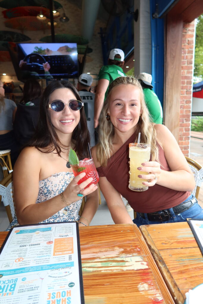 Two women smiling and holding colorful cocktails at Share House restaurant in Charleston, SC during a bachelorette party weekend planned by Bach Babes.