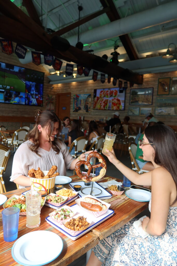 Two women toasting with cocktails over a spread of fried chicken, mac and cheese, a giant pretzel, and appetizers at Share House in Charleston, SC during a bachelorette party
