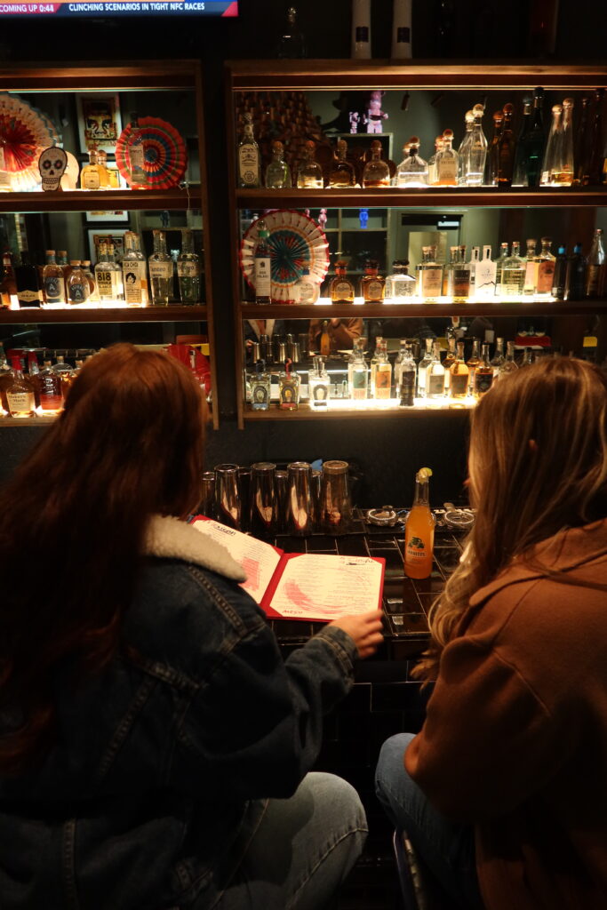 Two women sitting at the bar at Mesu restaurant in Charleston, SC, looking at the cocktail menu in front of an illuminated wall of tequila bottles during a bachelorette party outing.