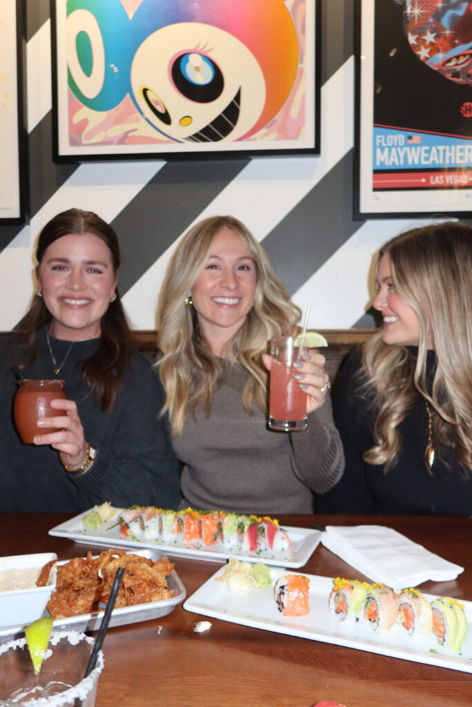 Three women smiling and holding cocktails in front of plates of colorful sushi rolls at Mesu restaurant on Upper King Street in Charleston, SC during a bachelorette party dinner planned by Bach Babes.