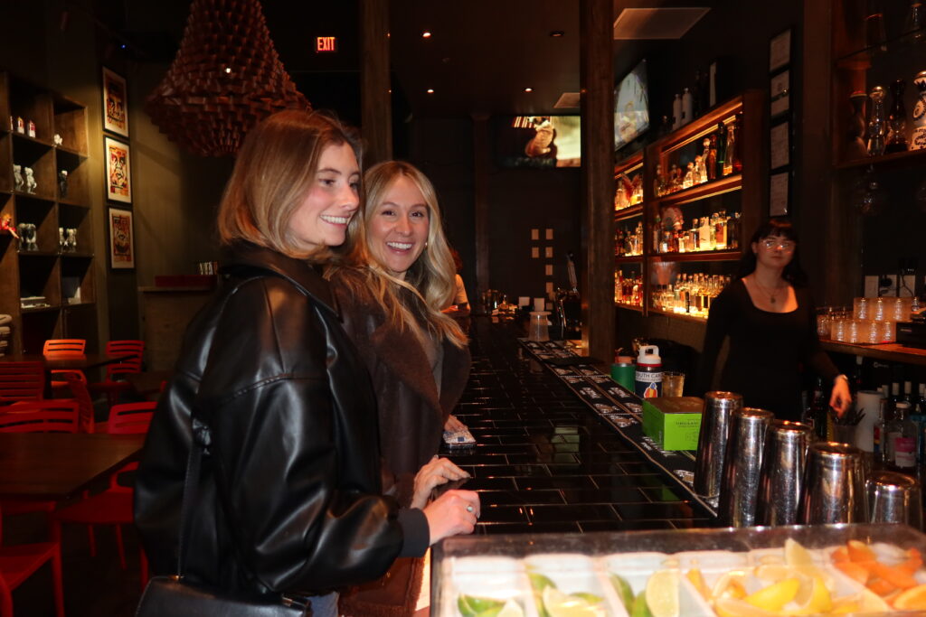 Two women smiling together at the bar inside Mesu restaurant in Charleston, SC, with an illuminated tequila bottle wall and bartender visible in the background during a bachelorette party night out.